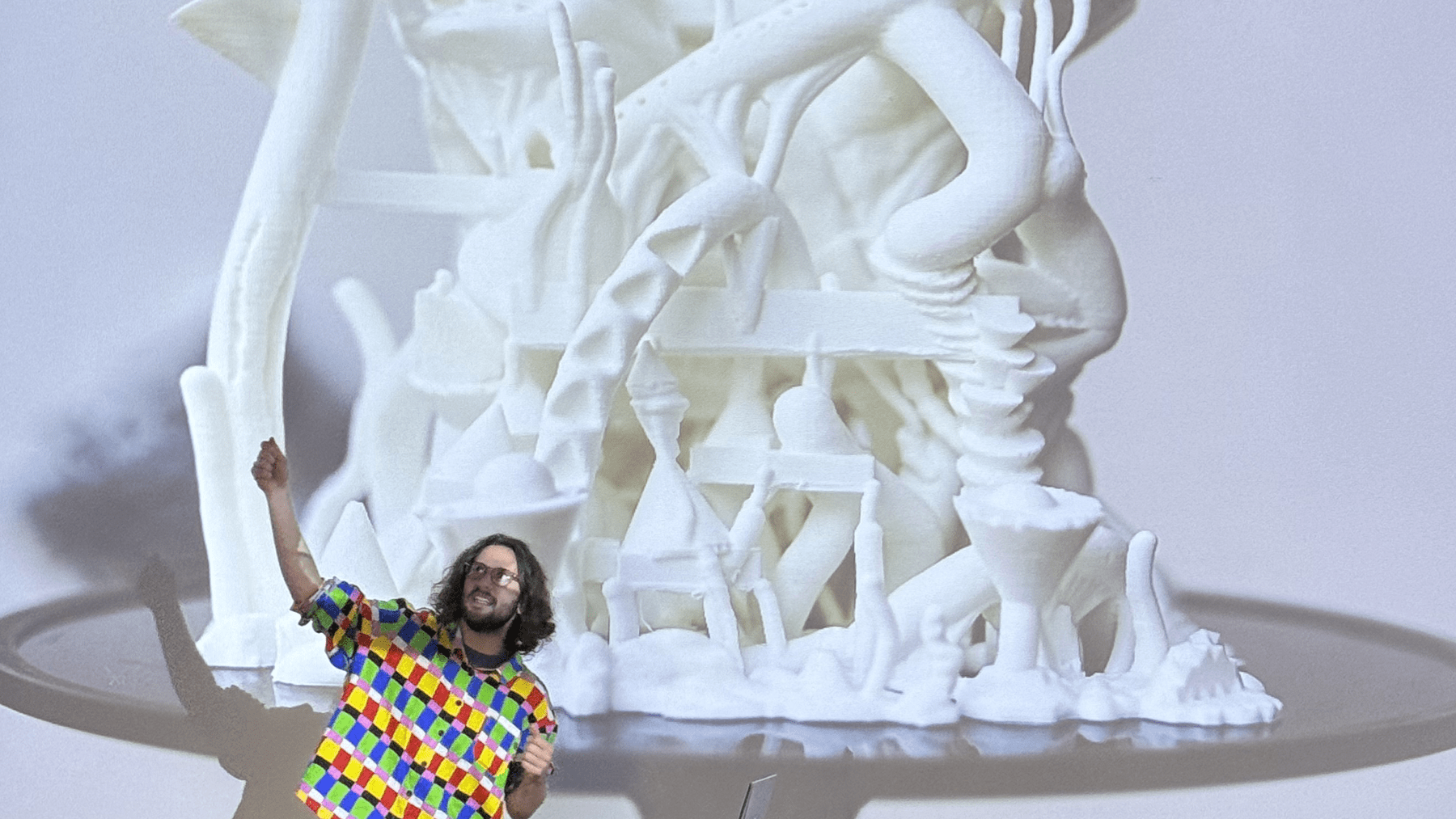 Shawn Chiki, in a brightly multicolored shirt, stands in front of a projected image of a digitally-sculpted architecture cast in white.