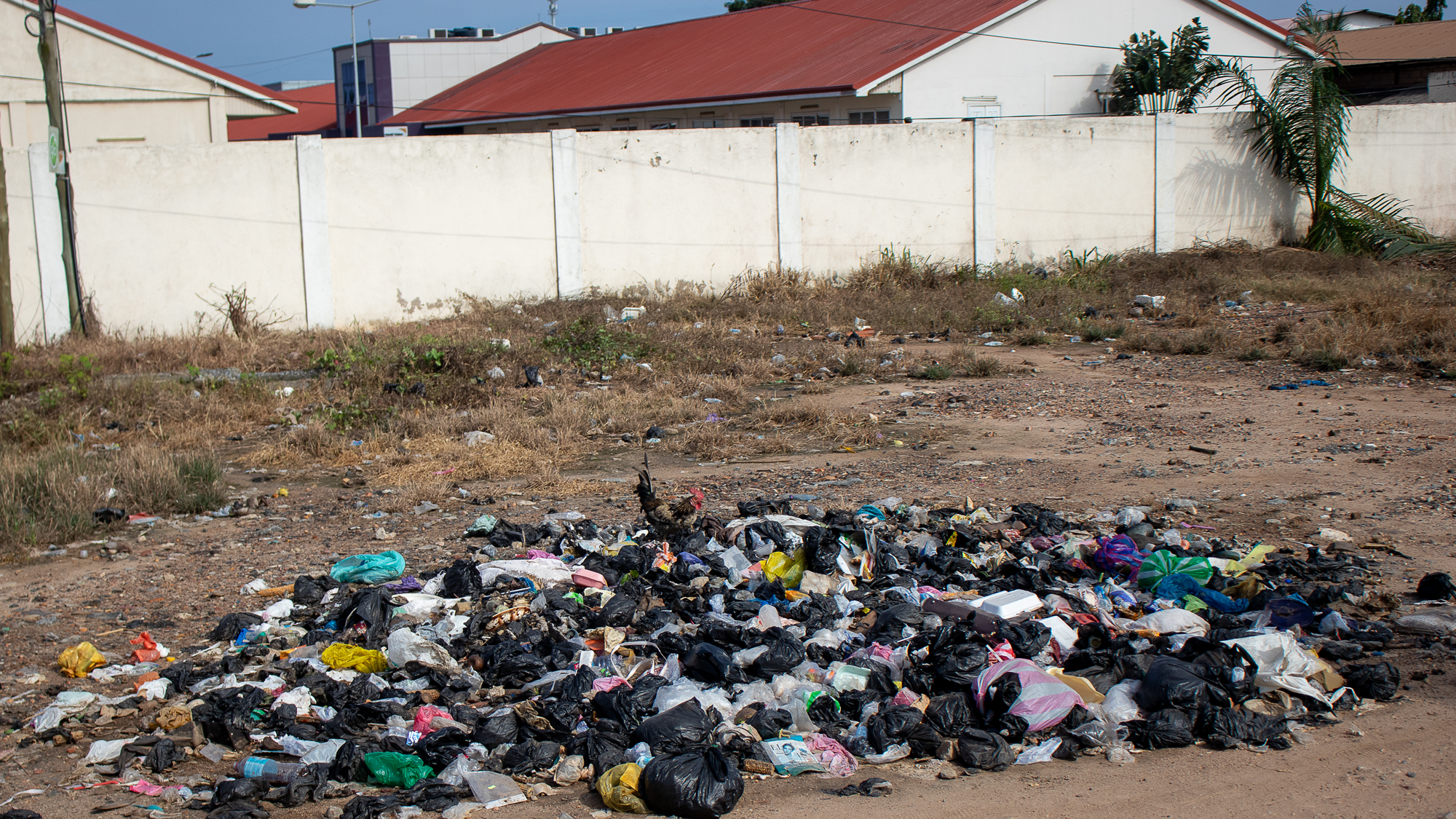 A pile of plastics waste on dirt ground with a white fence behind it, and red-colored roofs above the fence.