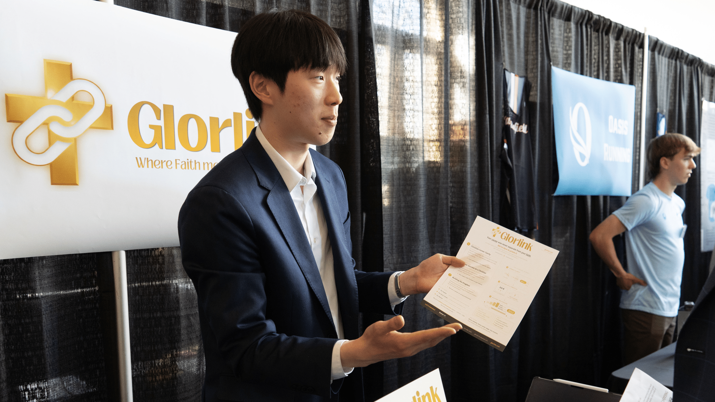A young East Asian man wearing a blue blazer and an open-collared white shirt holds up a printed "Glorlink" informational flyer with both hands while gesturing. He stands inside an event space with black backdrop curtains, flanked by a white "Glorlink" logo banner on the left and neighboring display booths on the right.