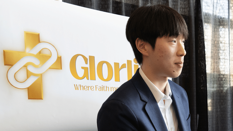 A young East Asian man wearing a dark blue blazer over an open-collared white shirt stands in profile on the right side of the frame, looking toward the right. Behind him to the left is a white banner featuring a gold cross with a white chain-link emblem and the text "Glorlink," set against a background of dark curtains.