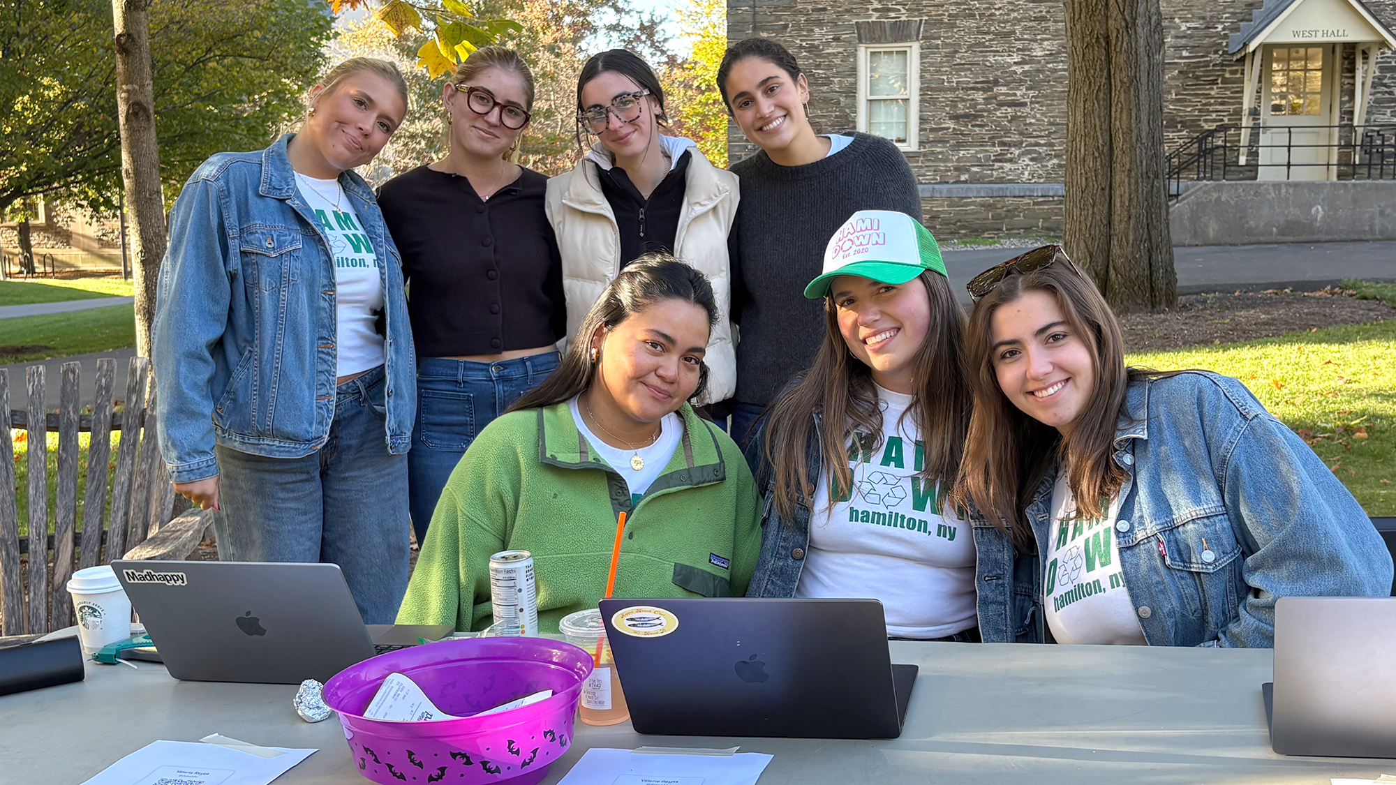 Four members of Hami-Down standing behind three members sitting at a table, mid-afternoon, wearing light jackets on a sunny day.