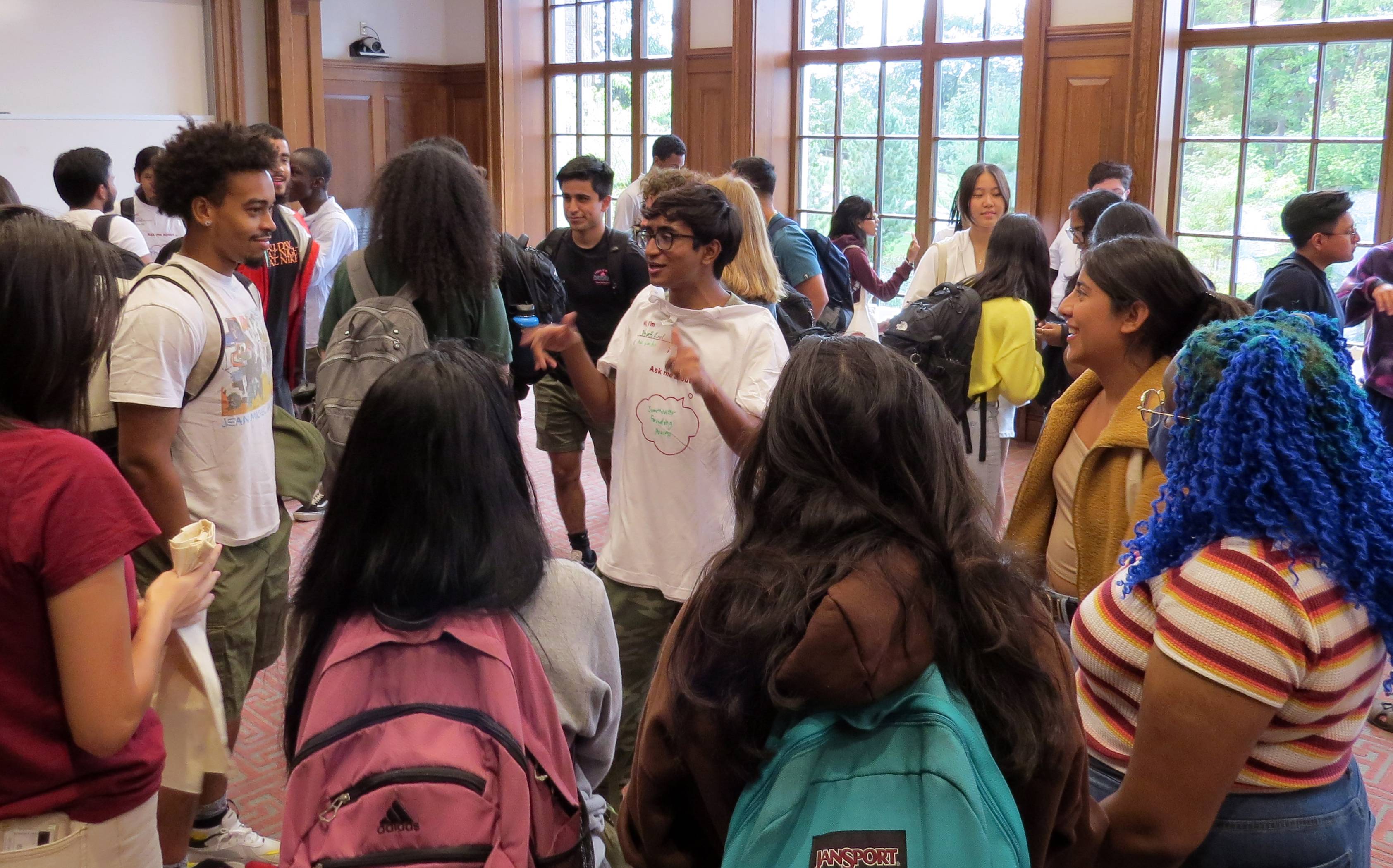 Students stand talking and laughing in group, wearing "Ask Me About" t-shirts.