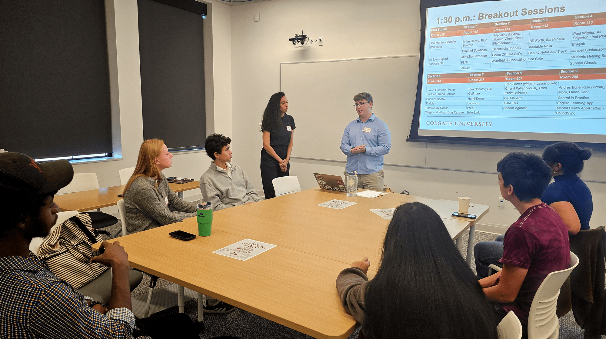 Students are seated around a table listening while a standing Geordie Waldman speaks in front of a projection screen.