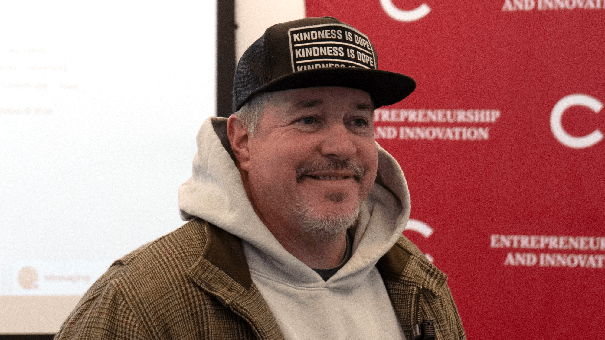 A man with a light beard and mustache smiles warmly while wearing a black hat featuring the repeated phrase “KINDNESS IS DOPE.” He is dressed in a grey hooded sweatshirt layered under a brown patterned jacket, standing in front of a red banner that reads “ENTREPRENEURSHIP AND INNOVATION.”