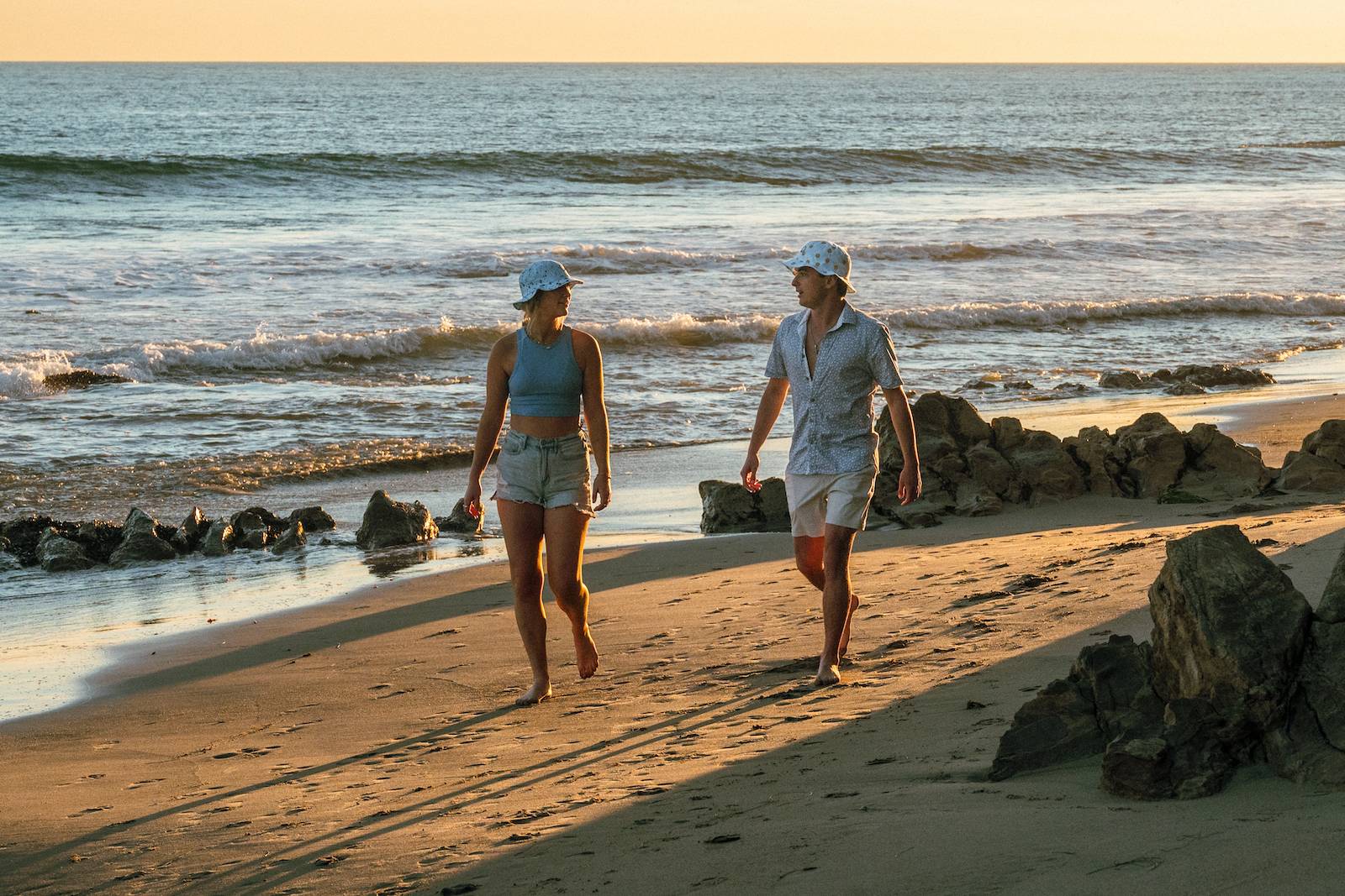 Two people walking on a sunny beach with bucket hats on.