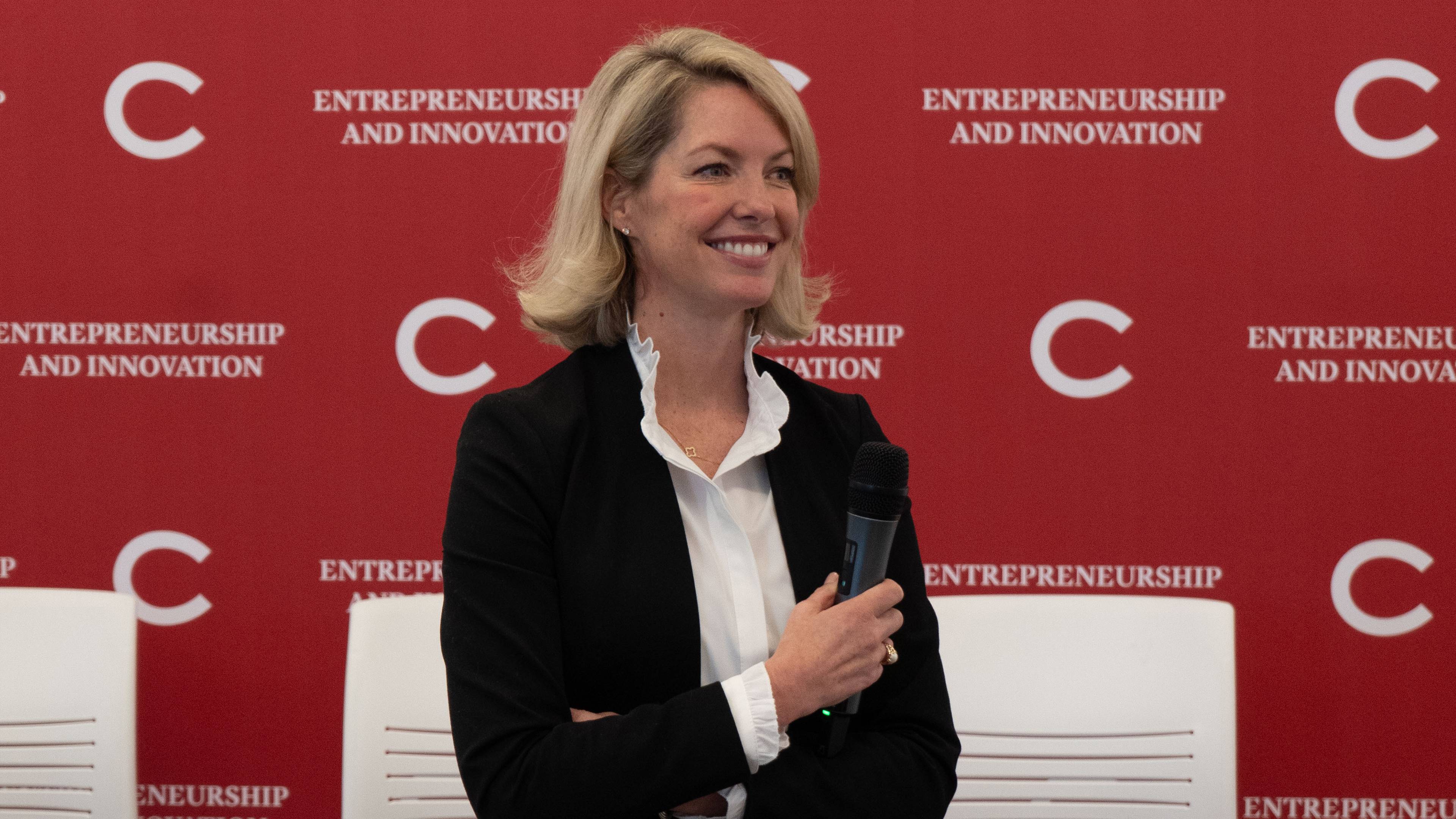 A smiling woman in a black blazer and white blouse holds a microphone in front of a red “Entrepreneurship and Innovation” backdrop. 