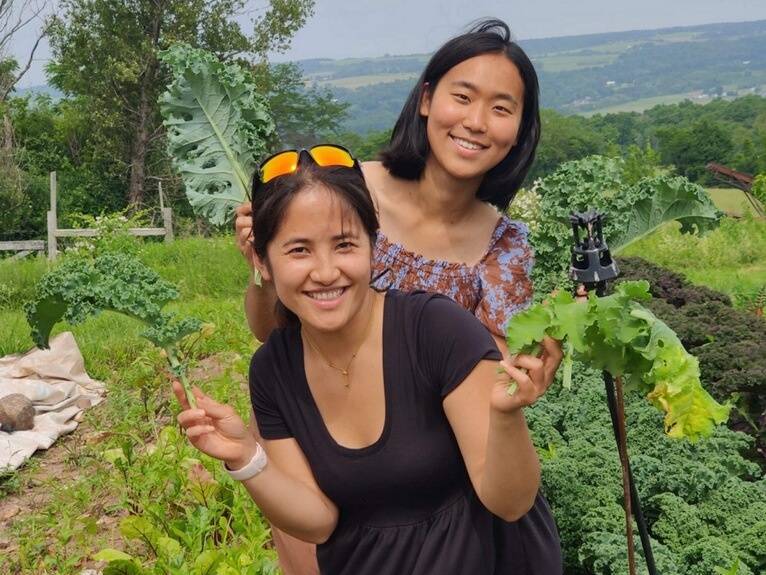 Sowon and Kay are holding kale leaves while standing in a field of vegetables at a local farm.