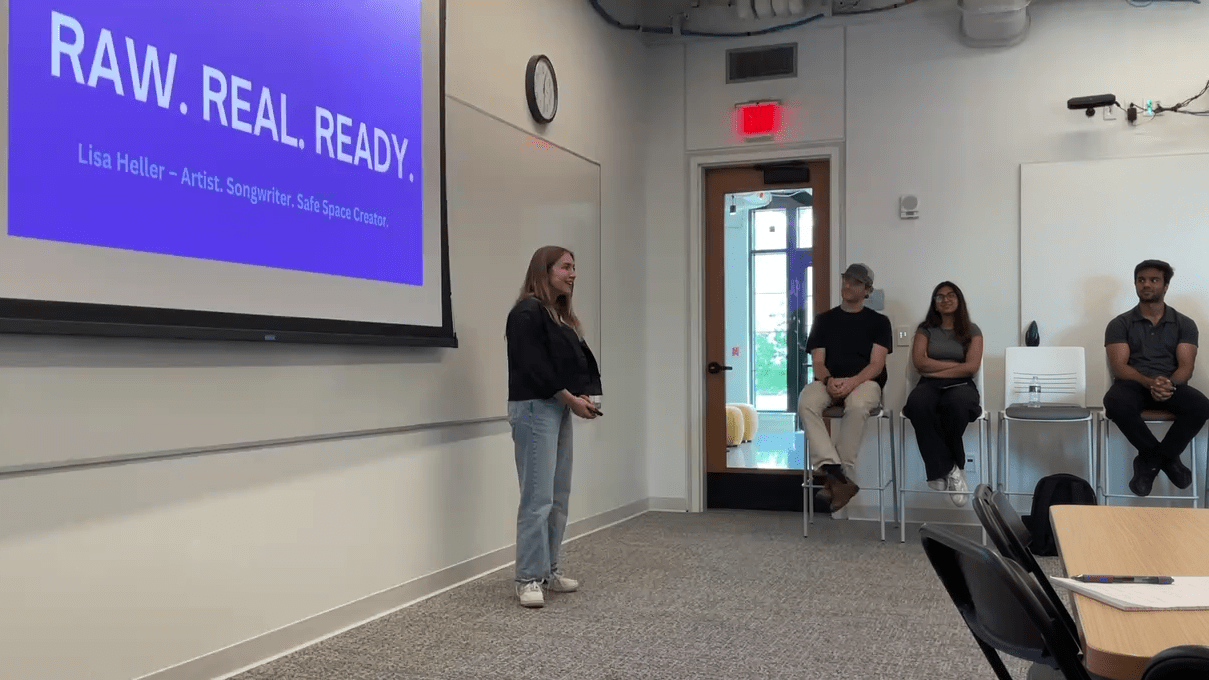 Lisa Heller standing at the front of a classroom speaking, with a PowerPoint presentation behind her that has the words raw, real, and ready displayed.