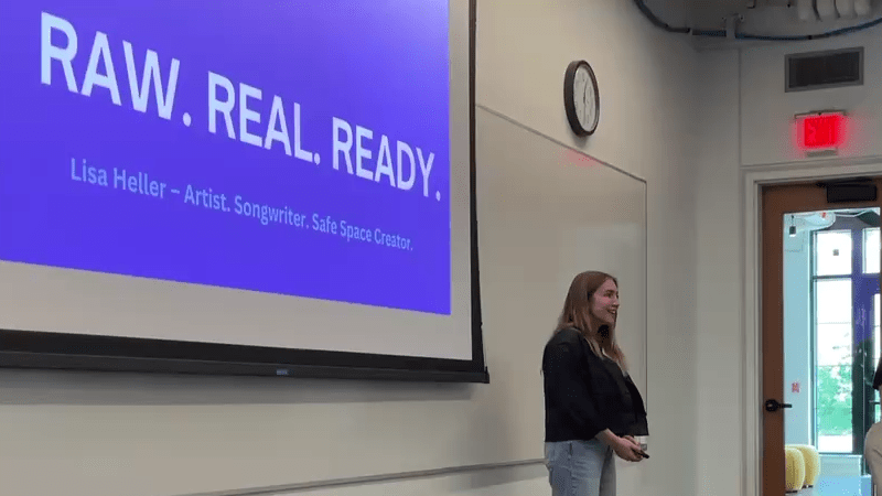 Lisa Heller standing at the front of a classroom speaking, with a PowerPoint presentation behind her that has the words raw, real, and ready displayed.