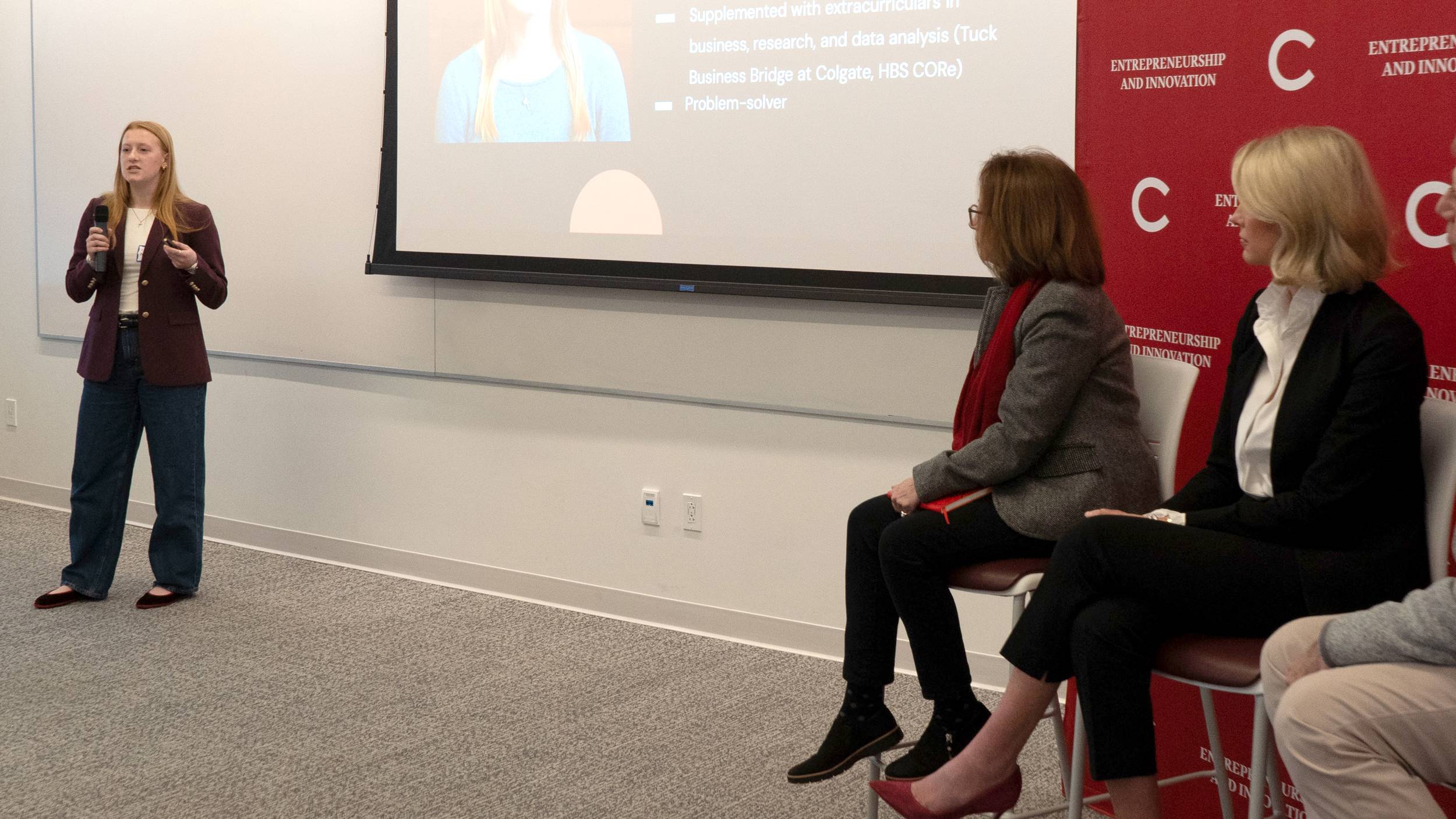 A young woman with long reddish-blonde hair stands on the left side of a classroom, holding a microphone and a clicker while presenting next to a large projection screen. On the right, two women sit on chairs watching the presentation in front of a red banner that reads "ENTREPRENEURSHIP AND INNOVATION" alongside a large letter "C".