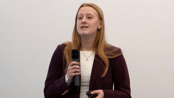 A medium close-up shot shows a young woman with long, straight reddish-blonde hair speaking into a handheld black microphone. She is wearing a maroon blazer over a white top, a simple necklace, and is holding a small black clicker in her other hand against a plain off-white background.