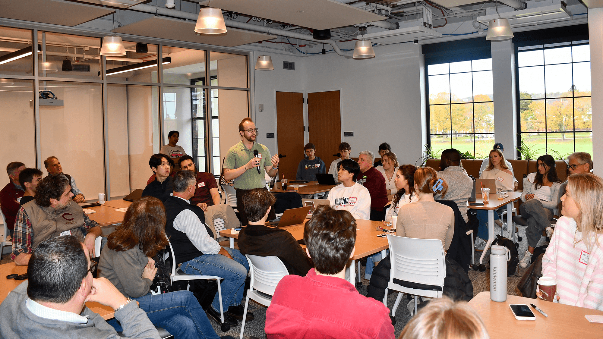 Mike Wilcox stands, speaking into a microphone in the center of a classroom surrounded by students seated and looking up at him.
