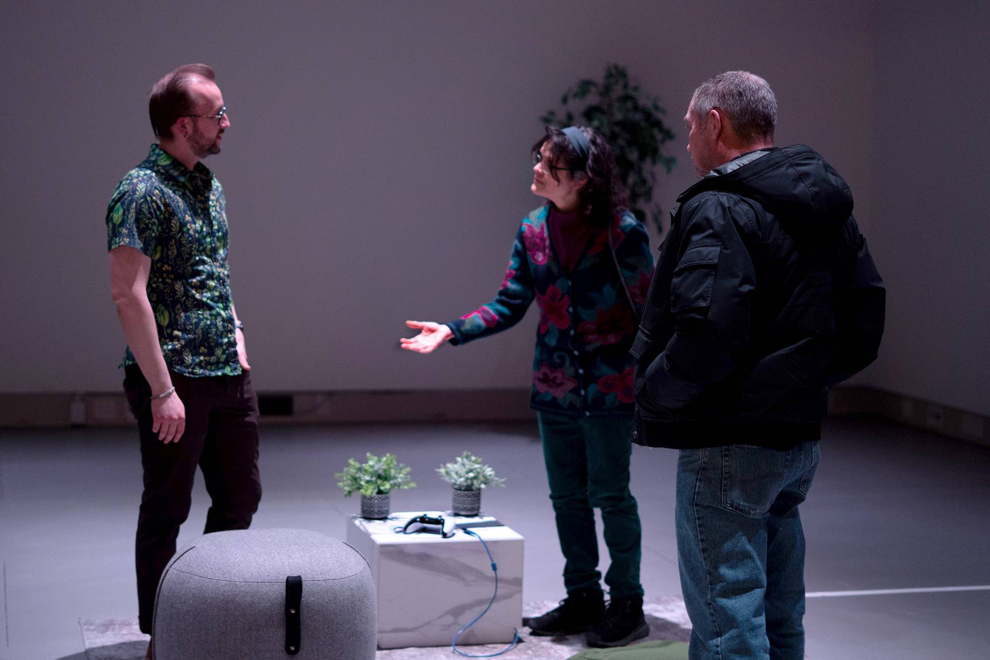Three people stand in a softly lit room having a conversation, with one person gesturing toward the others. A small table with two potted plants and a game controller sits between them. 