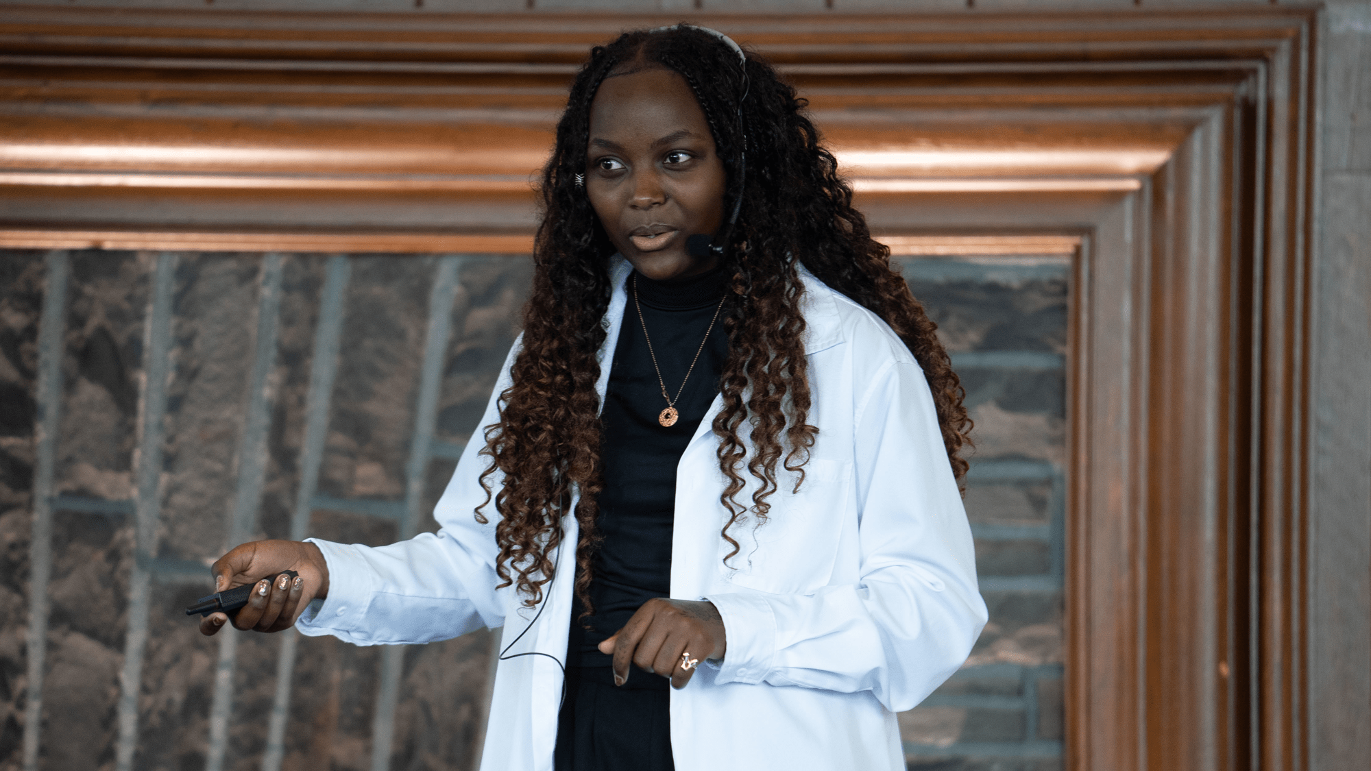 A speaker with long curly hair gestures while presenting, wearing a white button-down over a dark top and a headset microphone against a stone fireplace backdrop.
