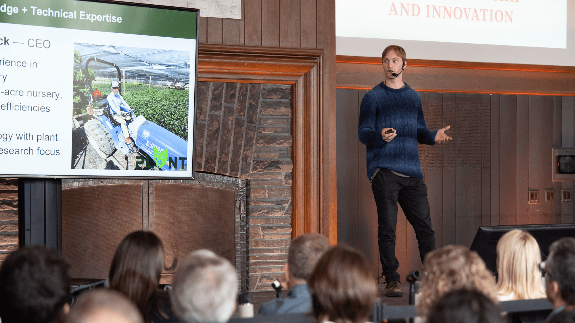 A young man in a blue sweater stands onstage with a headset microphone and clicker, presenting beside a large screen displaying a PLNT slide while audience members watch from the foreground.