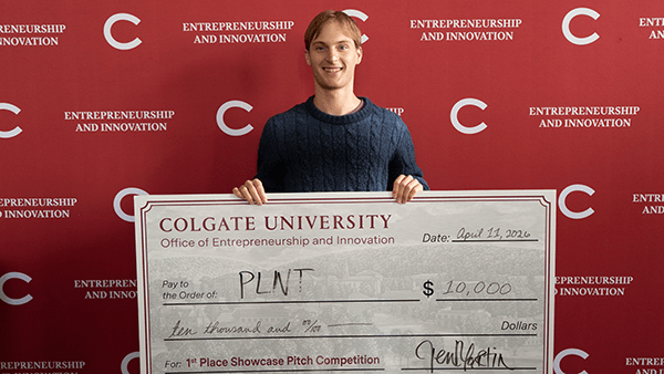 A smiling young man in a blue sweater stands in front of a red Entrepreneurship and Innovation backdrop holding an oversized check for $10,000 awarded to PLNT for first place in the showcase pitch competition.
