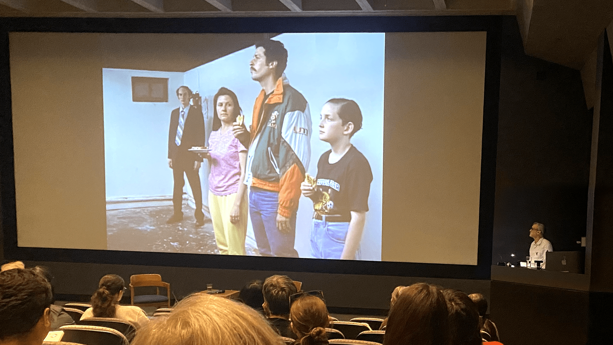 Richard Maxwell stands off to the side of a large movie screen, which has four characters standing in a row.