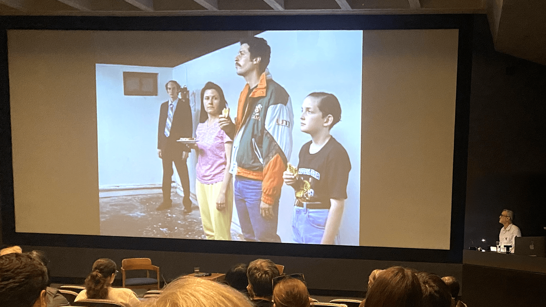 Richard Maxwell stands off to the side of a large movie screen, which has four characters standing in a row.