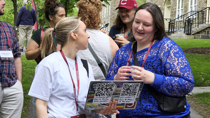 Outdoors on a paved walkway, a blonde woman wearing a white t-shirt and Colgate lanyard holds open a laptop toward another woman in a bright blue lace top. The back of the laptop displays stickers and two large QR codes with handwritten notes inviting viewers to scan for a demo and updates.