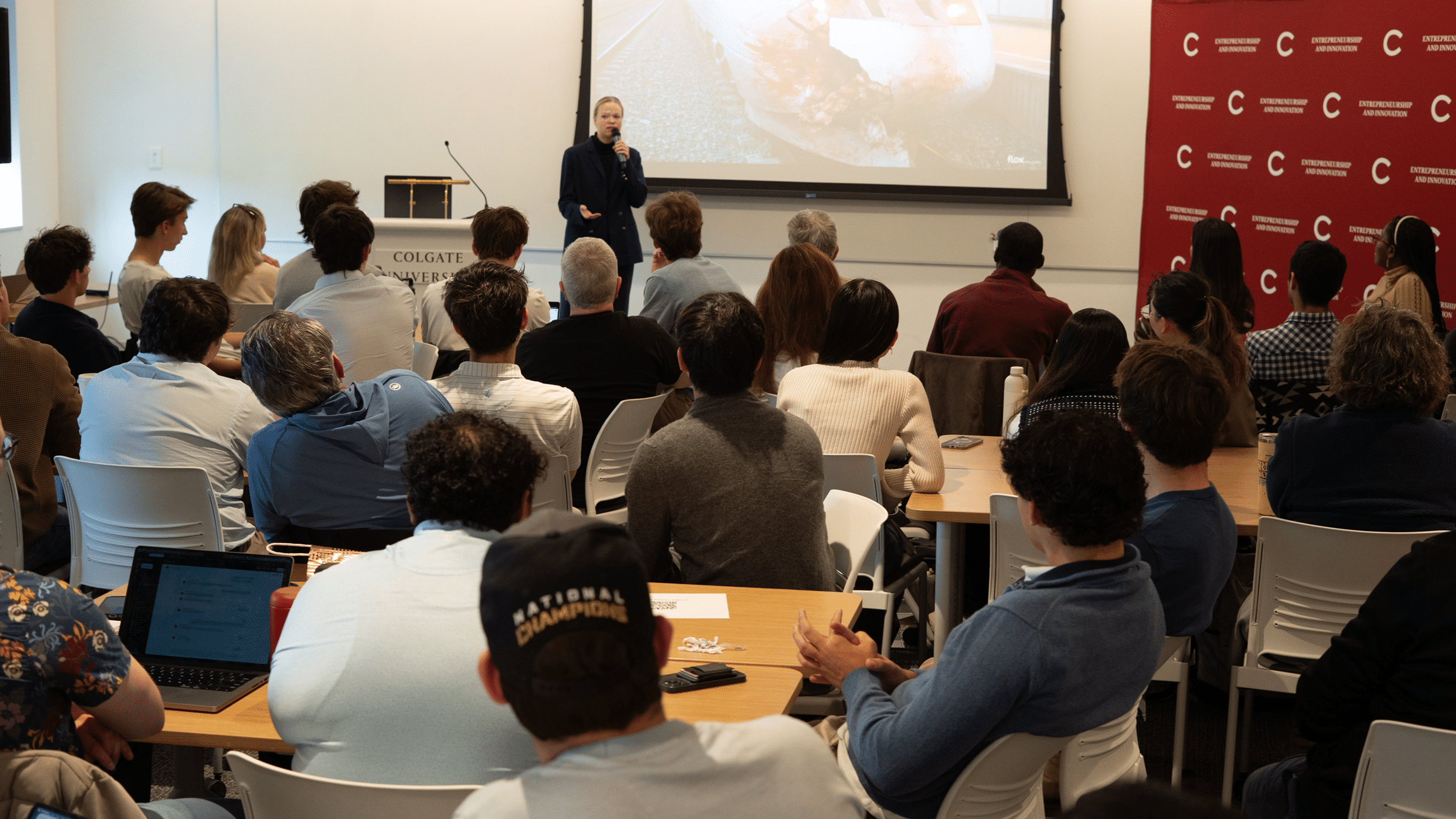 A speaker gives a presentation to a seated audience in a classroom-style event space.