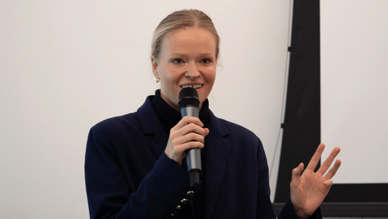 A woman speaks into a microphone while gesturing during a presentation.