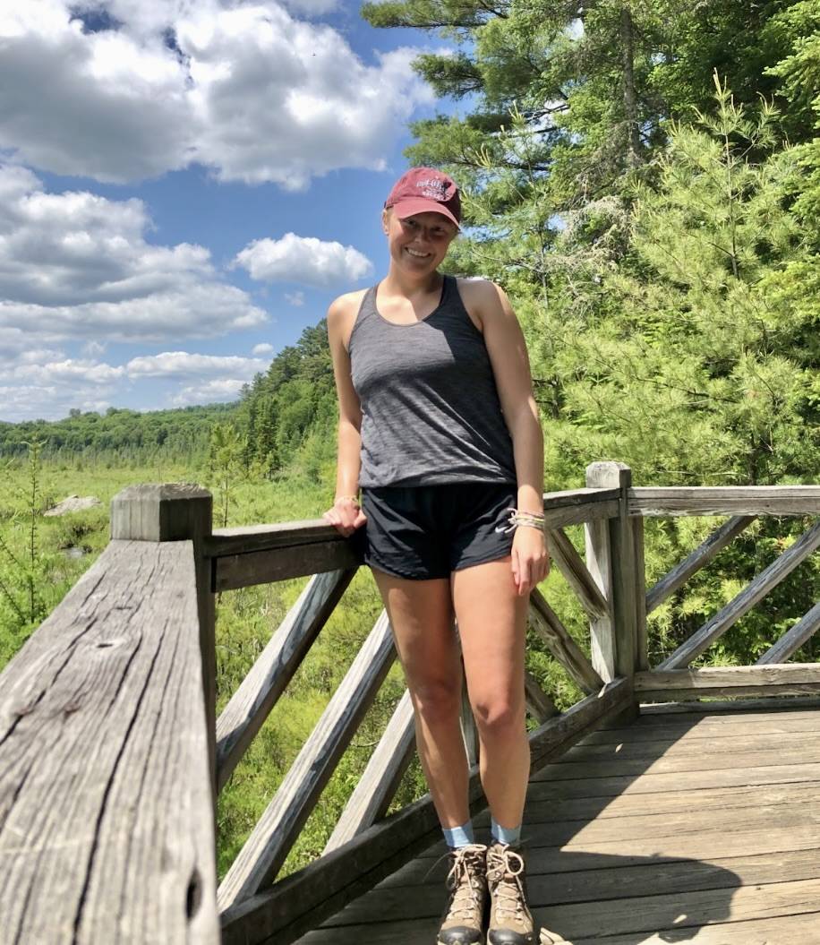 A photo of Kayla Gutheil standing on a wooden boardwalk with blue skies and trees behind her.
