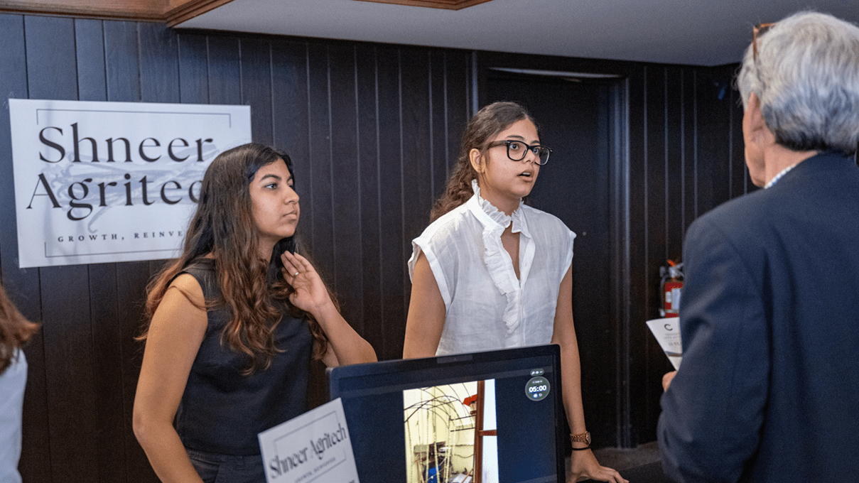 Colgate alumni Diya Badola and Jahanvi Chamria stand behind a table during ACI Weekend speaking with a member of the public about their idea.