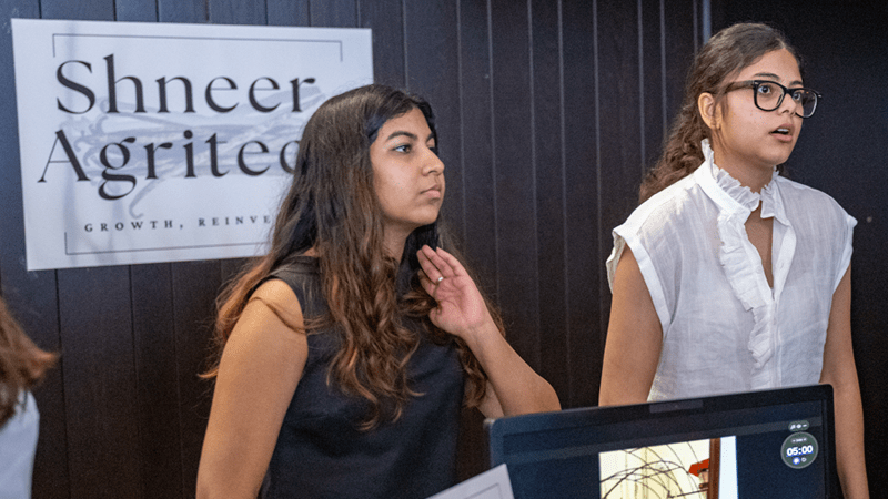 Colgate alumni Diya Badola and Jahanvi Chamria stand behind a table during ACI Weekend speaking with a member of the public about their idea.