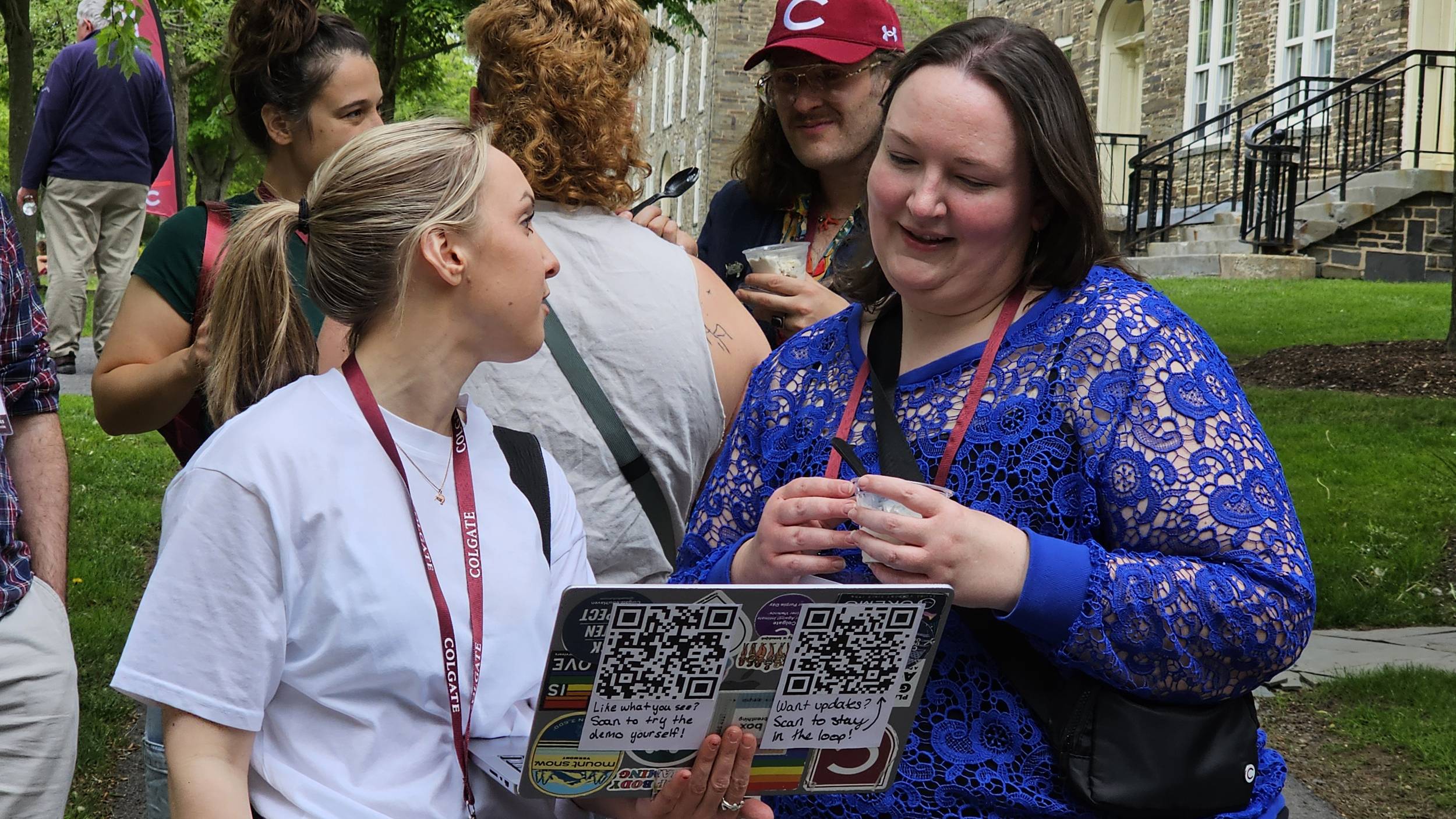 Outdoors on a paved walkway, a blonde woman wearing a white t-shirt and Colgate lanyard holds open a laptop toward another woman in a bright blue lace top. The back of the laptop displays stickers and two large QR codes with handwritten notes inviting viewers to scan for a demo and updates.