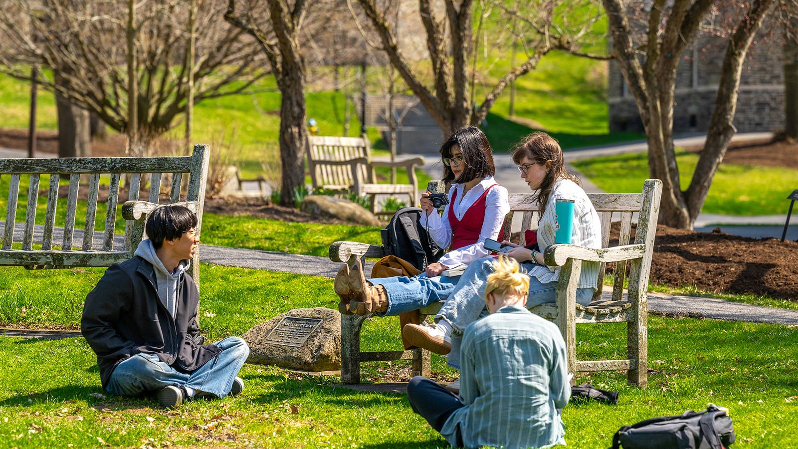 Students sit on a bench and on the grass on Colgate's campus while visiting on a sunny day. 