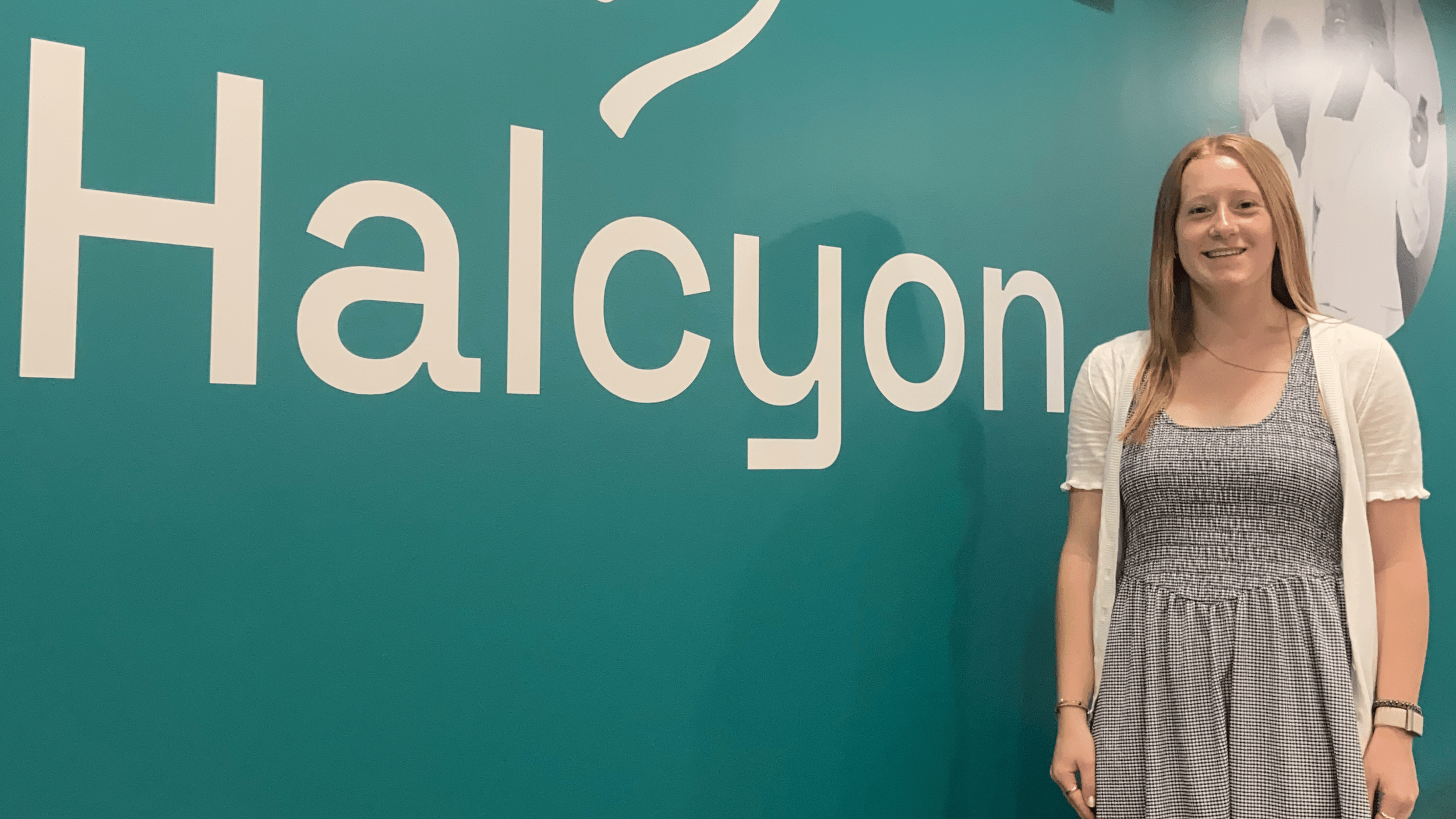 A smiling young woman with long blonde hair stands on the right side of the frame in front of a teal wall. Large white lettering on the wall behind her reads "Halcyon" alongside part of a logo above it.