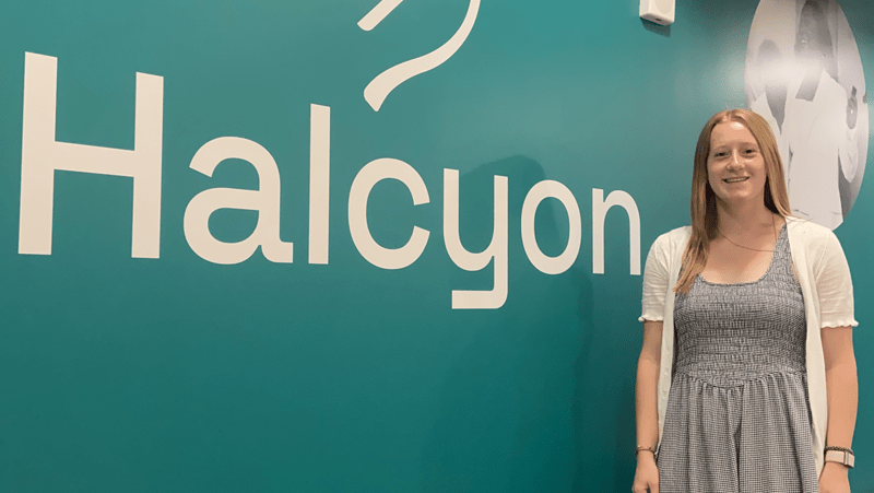 A smiling young woman with long blonde hair stands on the right side of the frame in front of a teal wall. Large white lettering on the wall behind her reads "Halcyon" alongside part of a logo above it.