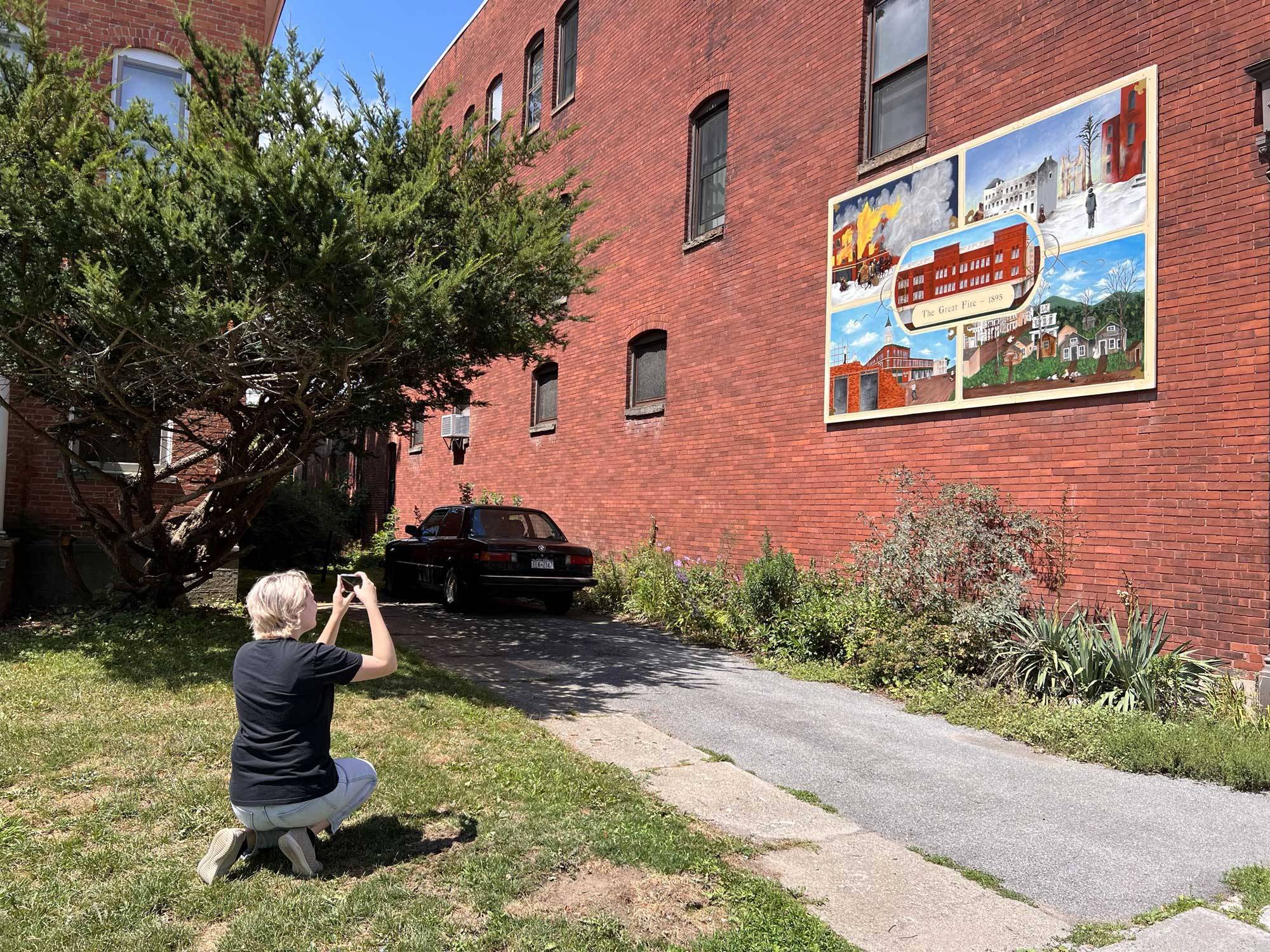 Rebecca Sweigart is kneeling in front of a mural that depicts the history of the Chenango Canal to video the image for her documentary.