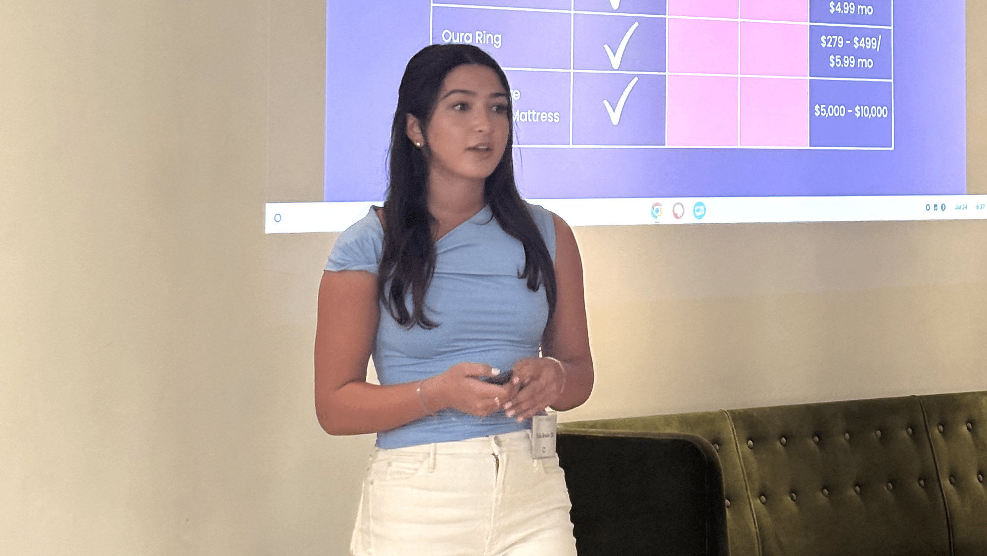 Talia Broder stands at the front of a room with a slide presentation on the wall behind her, comparing her product to others.
