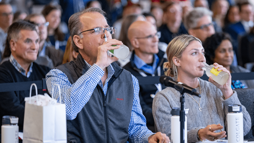 A man in a grey vest and a woman in a grey sweater sit at a table in a crowded auditorium, both simultaneously drinking from small glass bottles containing a pale liquid. They are judges participating in the Entrepreneurship Showcase, with white Colgate-branded water bottles and a microphone positioned on the table in front of them.
