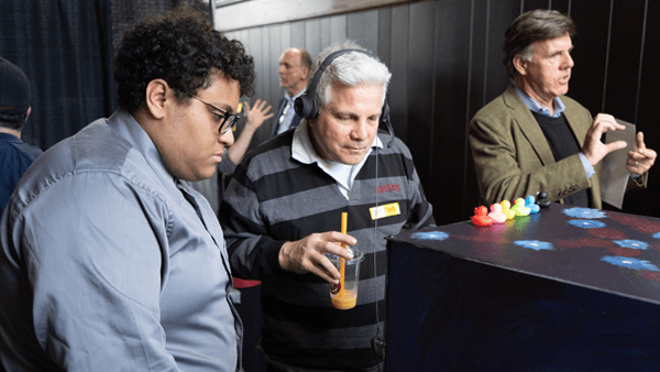 A younger man with glasses and a gray dress shirt looks down intently alongside an older man wearing a headset and a striped sweater who is holding a drink with a straw. They are gathered around a dark table featuring a row of small, colorful rubber ducks, while another man in a blazer gestures in the background.