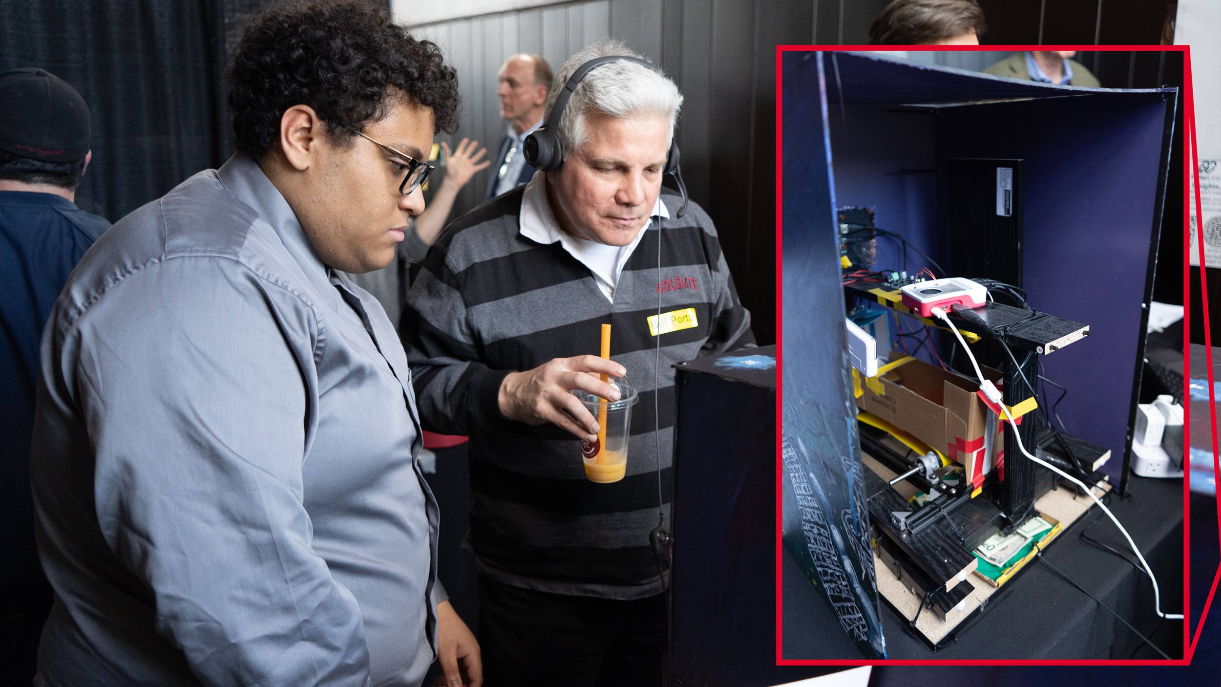 A young man wearing glasses and a gray shirt looks down alongside an older man in a headset and striped sweater holding a beverage. A large inset photo with a red border highlights the interior of the dark box they are examining, revealing a complex electronics setup with wires, a small computer board, a cardboard structure, and mechanical parts.