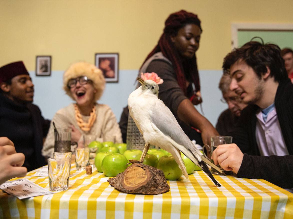 Cast sitting around a table with a yellow and white plaid tablecloth with a seagull sitting in top of the table. 