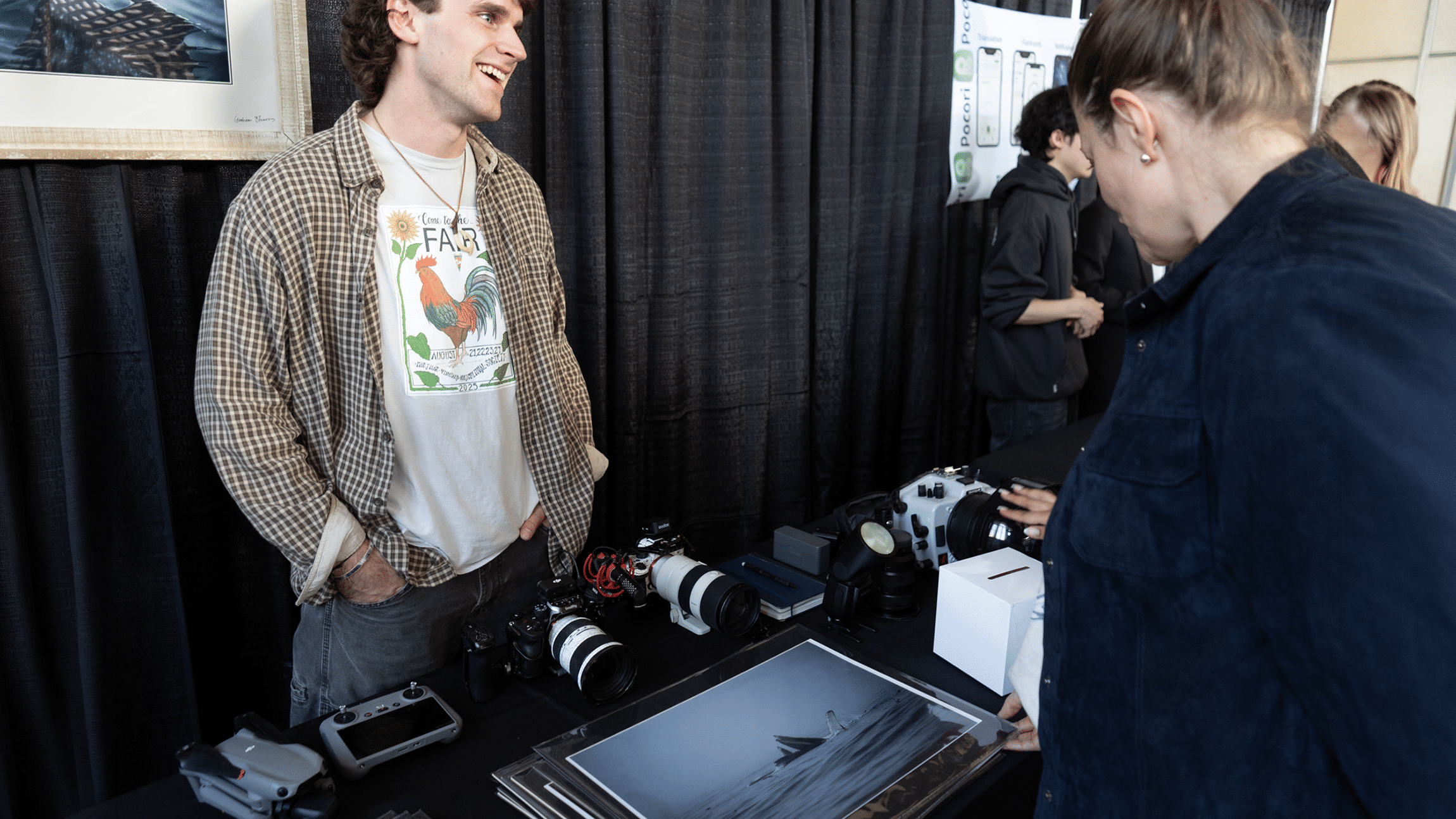 A smiling young man wearing an open plaid shirt over a graphic t-shirt stands behind a display table, interacting with a visitor in the foreground. The table is filled with photography gear and artwork, including professional cameras with telephoto lenses, a drone and controller, photo prints, and an underwater camera housing against a black curtain backdrop.