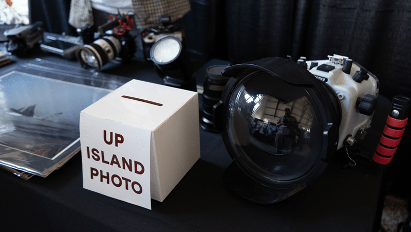 A white card box with a slot on top labeled "UP ISLAND PHOTO" sits on a dark display table next to a large underwater camera housing with a prominent curved dome lens. Additional photography equipment, including various camera lenses, a flash unit, and packaged photo prints, rests in the background against a dark curtain.