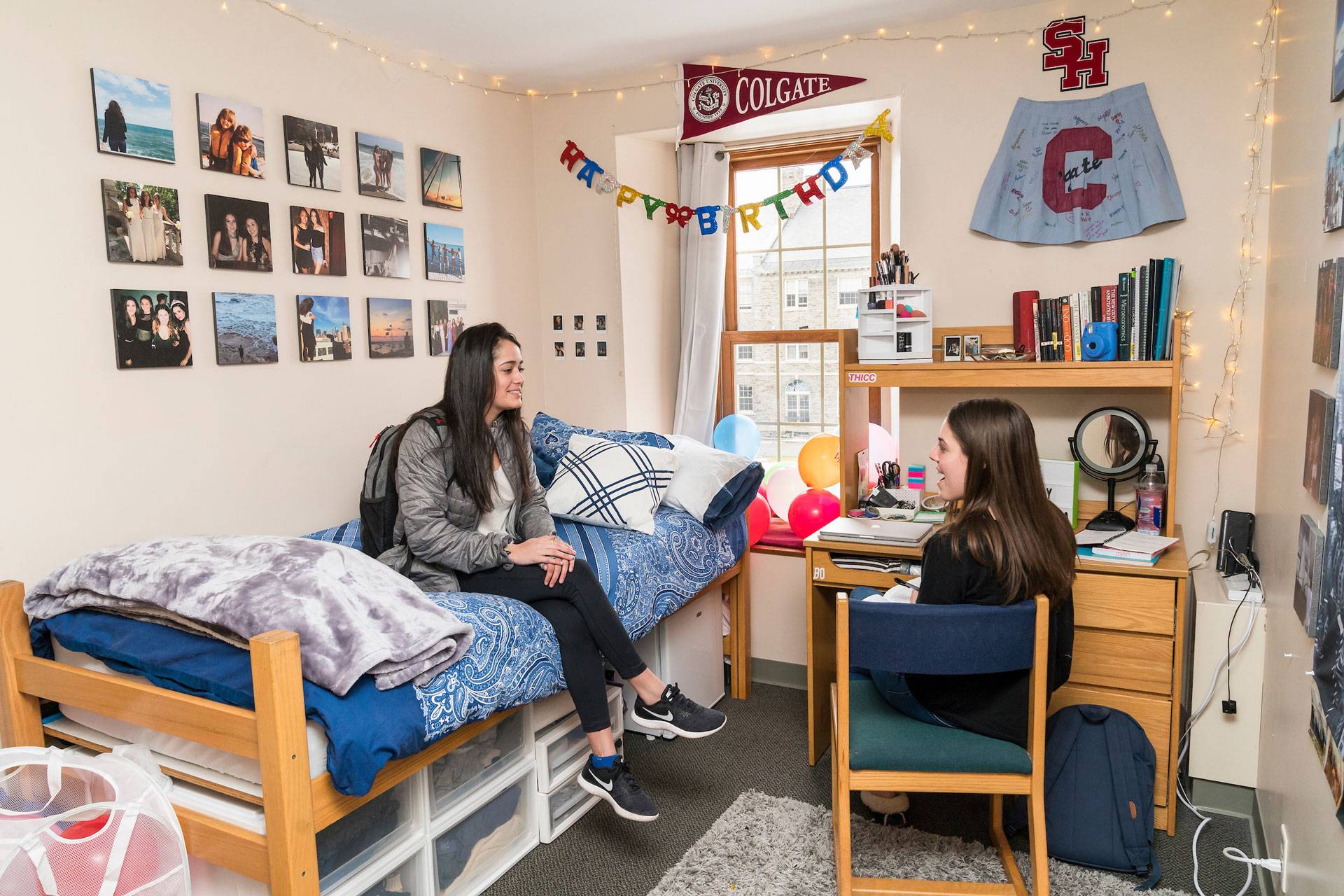 Two women, both with long, dark hair, socialize in a West Hall room. One sits on the bed, the other in a chair at the desk next to the bed. The walls are decorated with a Colgate pennant, Happy Birthday banner, and a grid of personal photos.