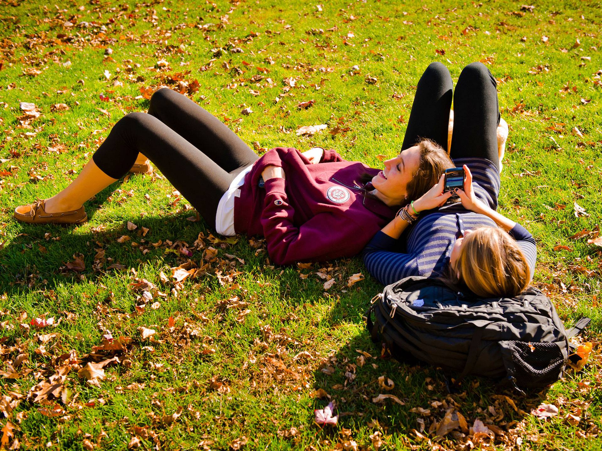 Two female students lying on grass with autumn leaves around them