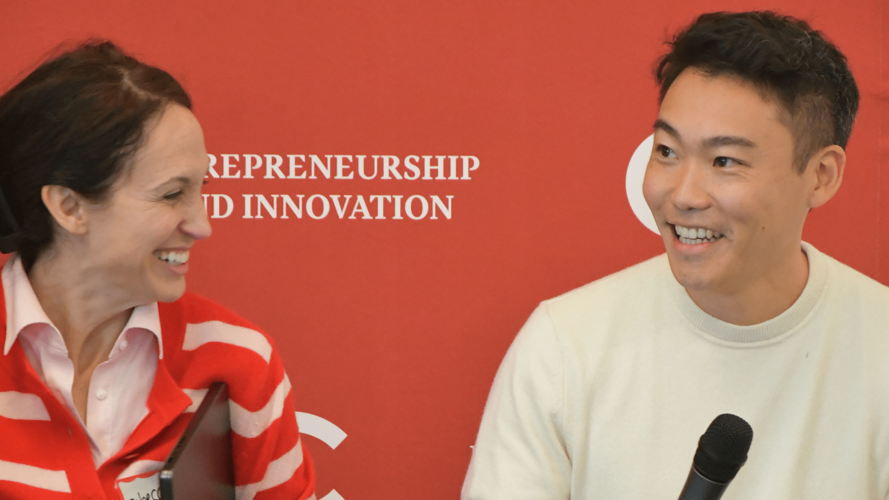 Rebecca Nackson and Yuni Baker-Saito seated, holding microphones, and laughing hard.