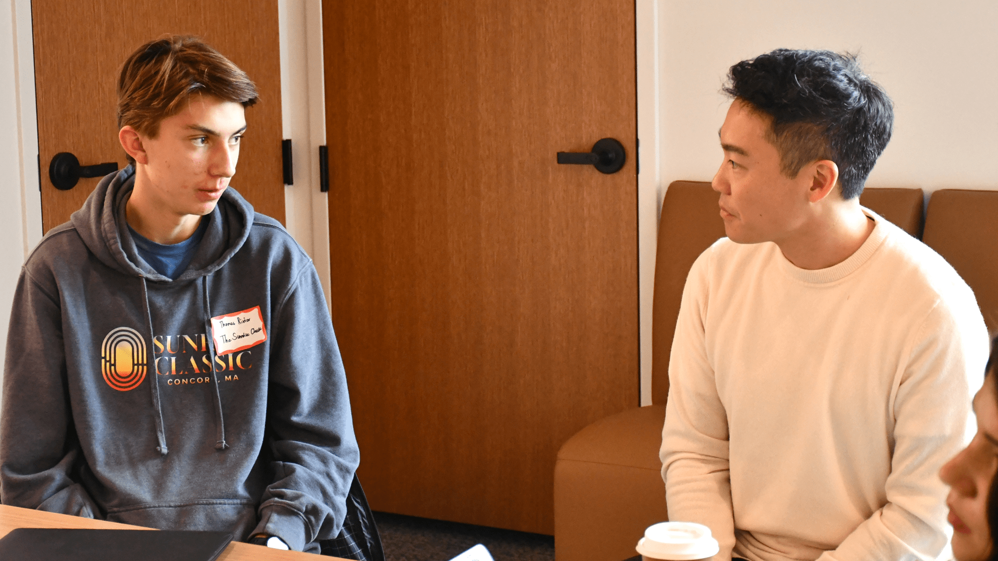 Thomas Rivier and Yuni Baker-Saito are seated at a table, looking toward each other and talking.