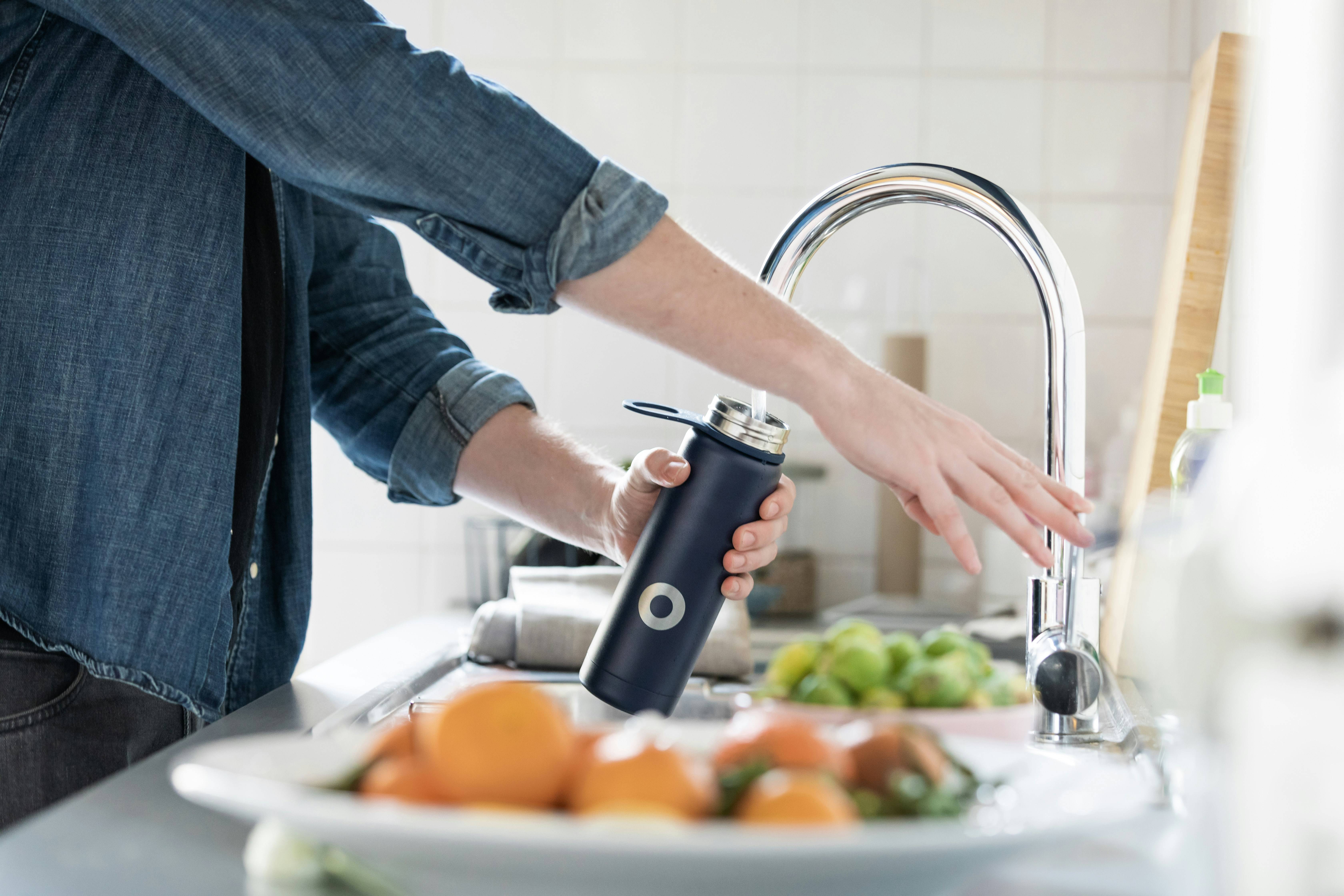 An arm extends to hold a water bottle under a running kitchen faucet. 