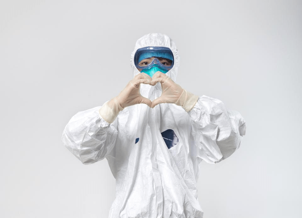 Photo description: A person in a hazmat suit, arms at shoulder height, with hands and fingers shaped in a heart