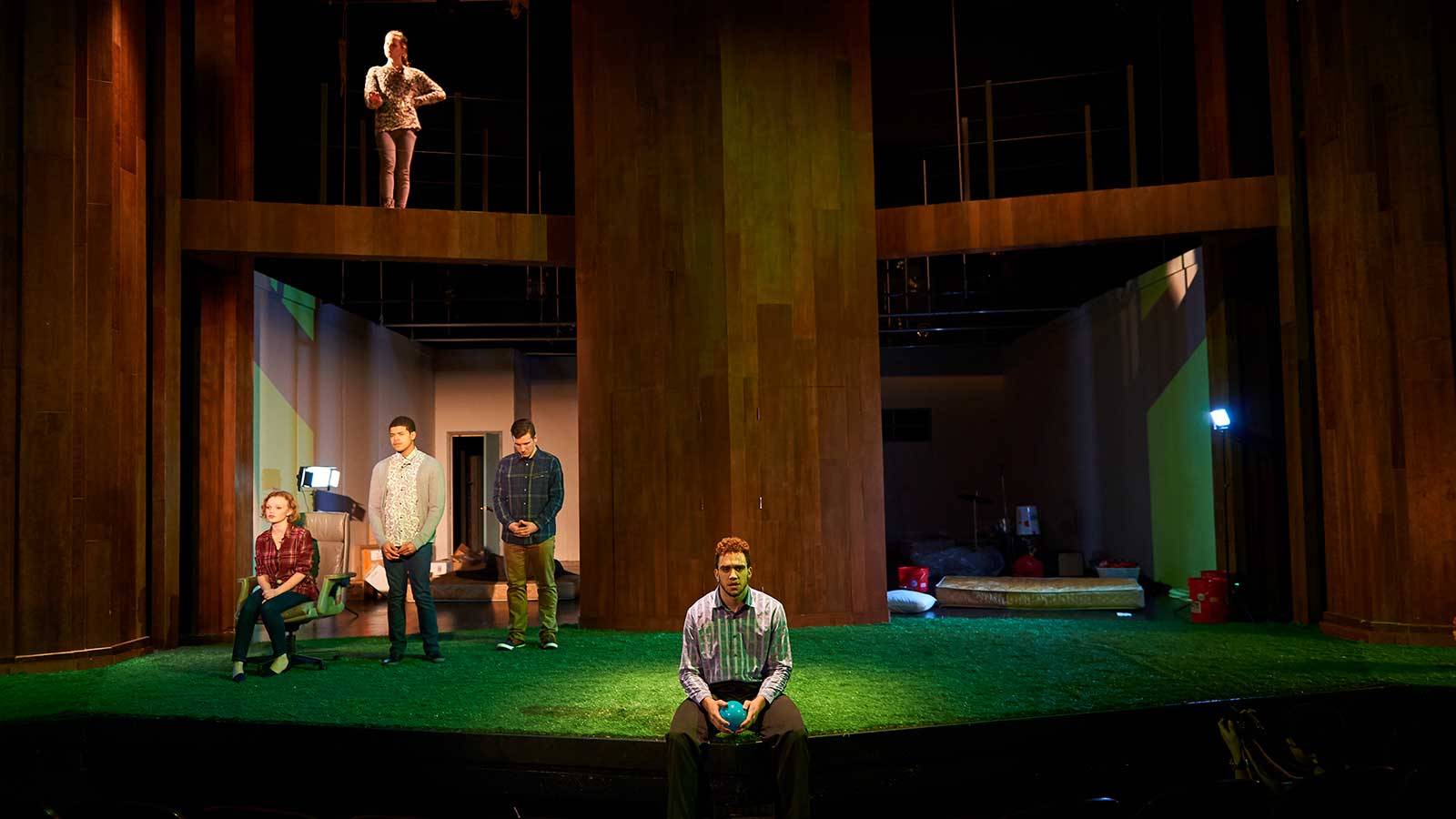 A performer sits on the edge of the stage with additional performers on two levels behind him on stage