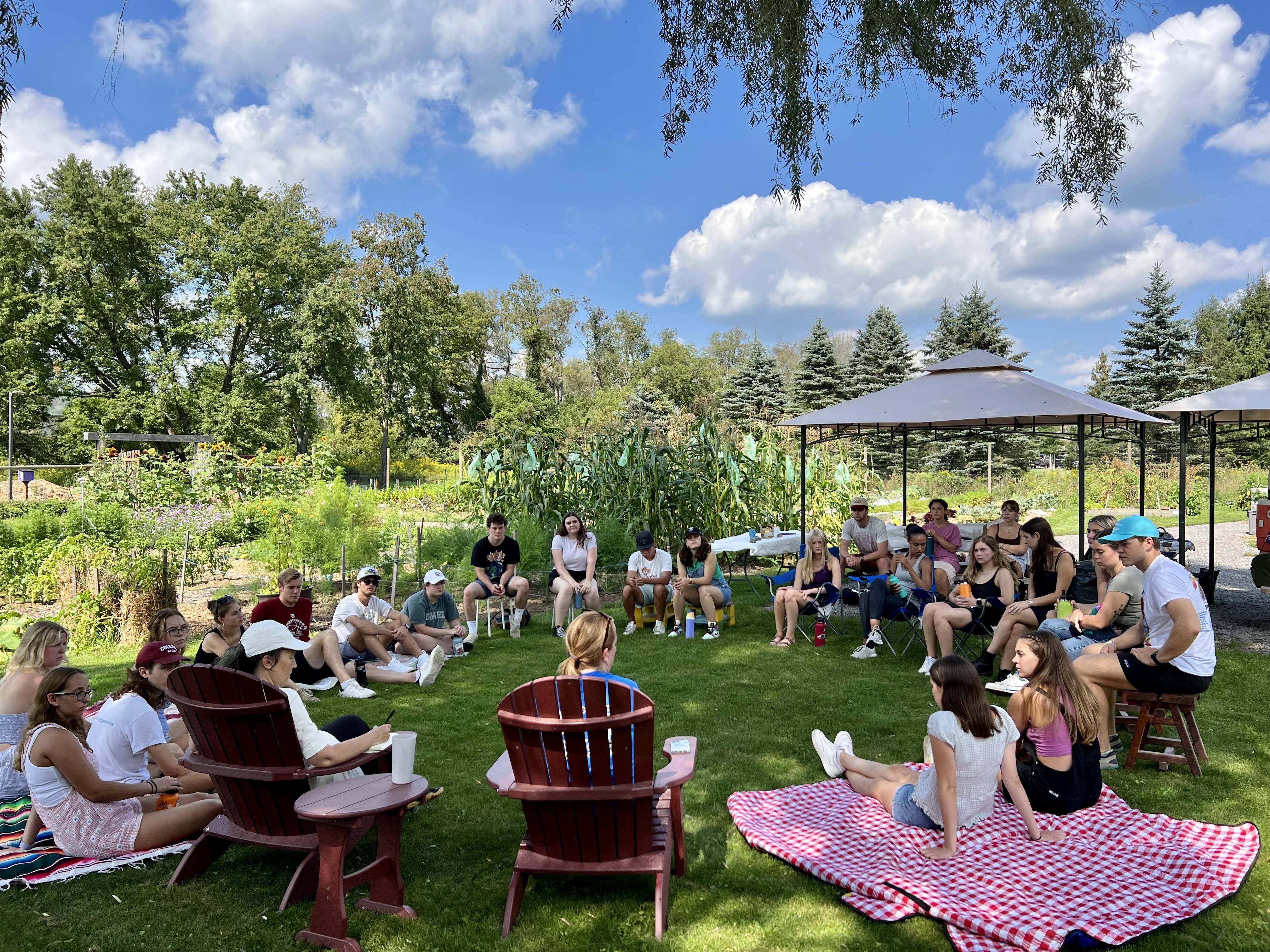 Photo of ENST 450 students sitting in a circle on the grass at the Community Garden under blue skies.