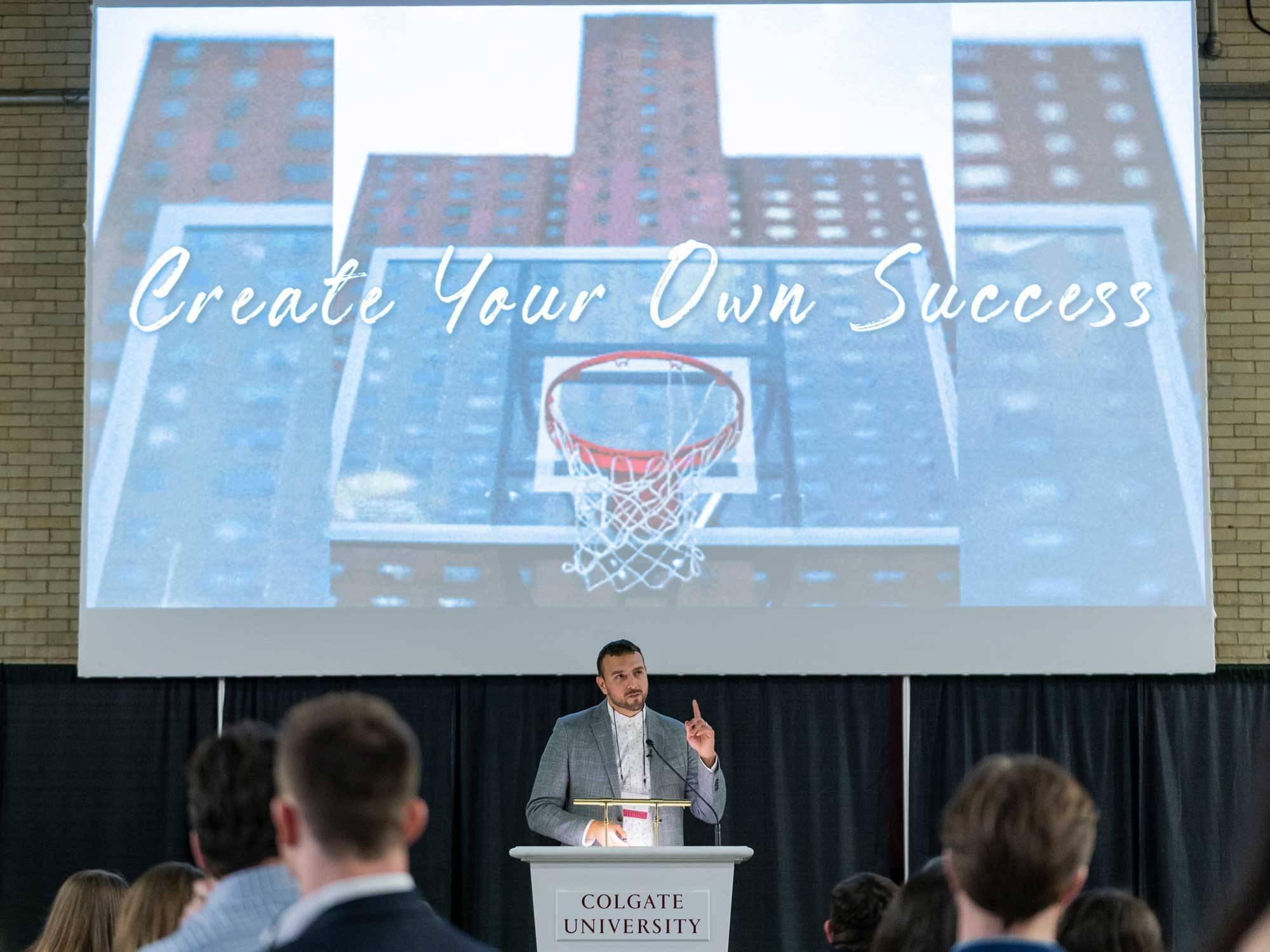 Keynote speaker Jon Lopez ’06 stands below picture of basketball hoop with caption "Create Your Own Success"