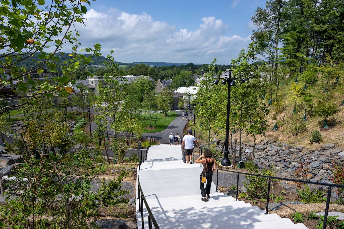 View of a person walking down Peter’s Glen in a landscaped area, with trees and a glimpse of a Colgate University in the background under a clear sky.